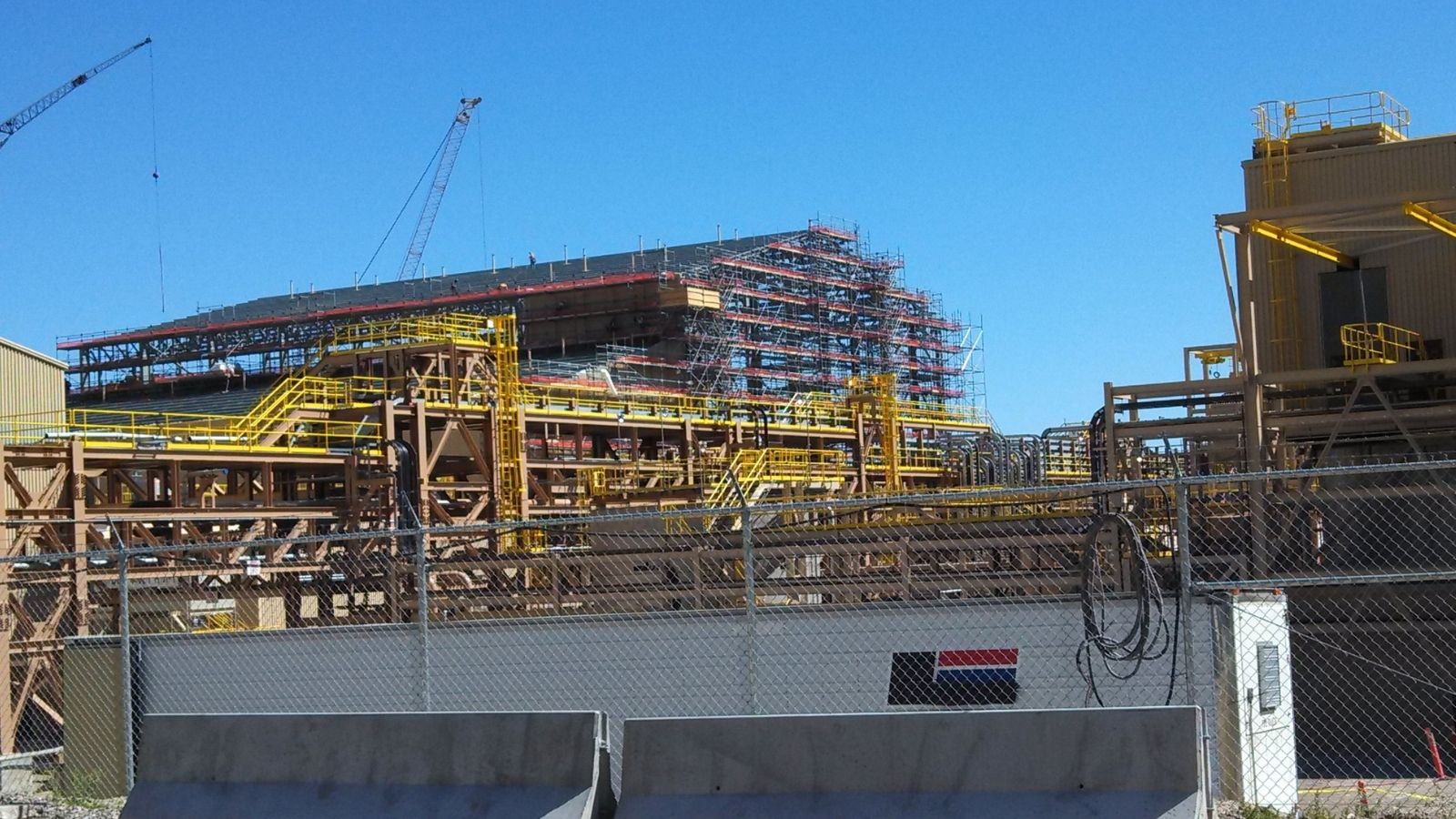 Industrial construction site with scaffolding, cranes, and metal structures behind a chain-link fence under a clear blue sky.