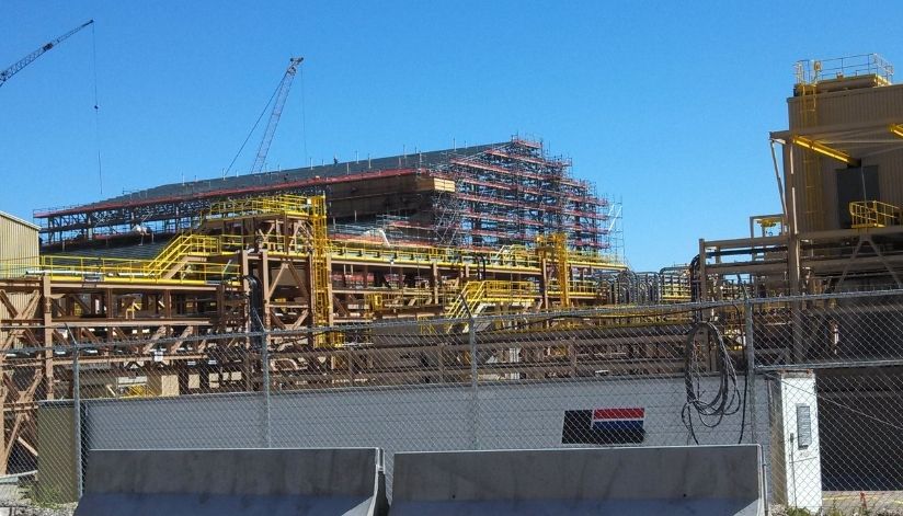 mid-city-steel-kennecott-magna-utah-industrial-build-fabrication-featured An industrial construction site with scaffolding, cranes, yellow railings, and fenced-off areas under a clear blue sky.
