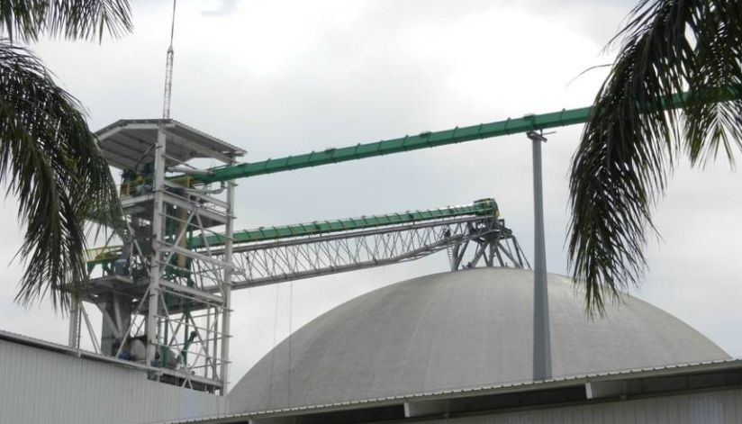 mid-city-steel-la-union-sugar-special-project-gallery-2 Industrial facility with a large dome, conveyor belts, and palm trees in the foreground under a cloudy sky.