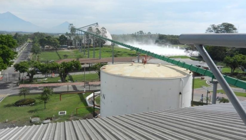 mid-city-steel-la-union-sugar-special-project-gallery-5 A large white industrial tank near a green conveyor belt with trees and mist in the background.