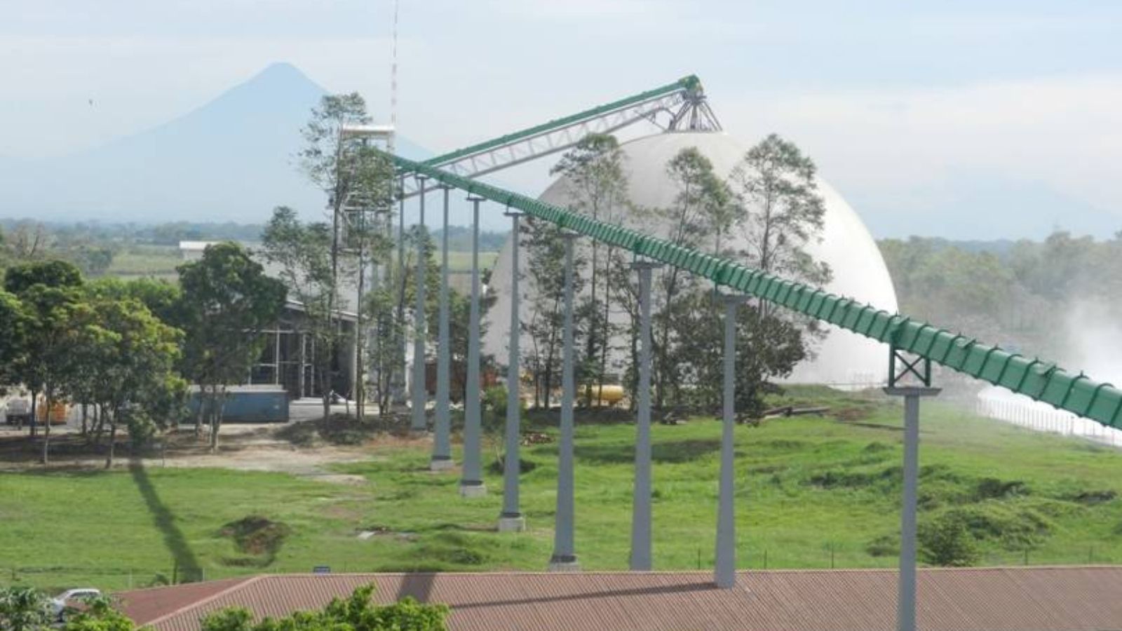 A large dome structure stands behind trees, with a green conveyor belt extending over a grassy area.