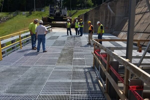 mid-city-steel-michigan-service-bridge-industrial-project-gallery-1 Several construction workers in safety gear stand on a metal grating platform next to a concrete wall outdoors.