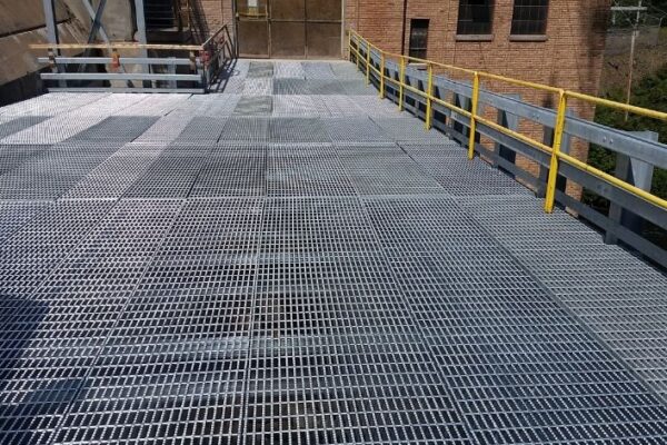 mid-city-steel-michigan-service-bridge-industrial-project-gallery-2 A metal grating walkway with yellow railings beside an industrial brick building.