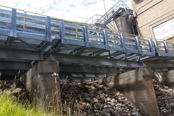 mid-city-steel-michigan-service-bridge-industrial-project-gallery-3 A metal pedestrian bridge on concrete supports spans over rocks beside a brick industrial building.