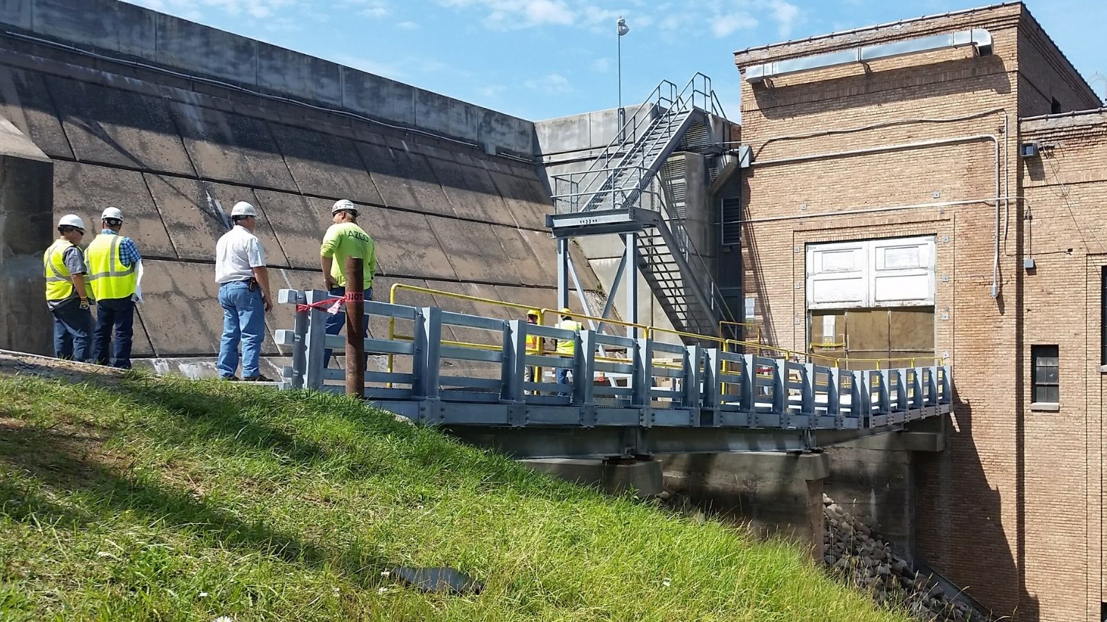 Several workers in safety gear stand on a walkway near a brick industrial building and concrete structure under a blue sky.