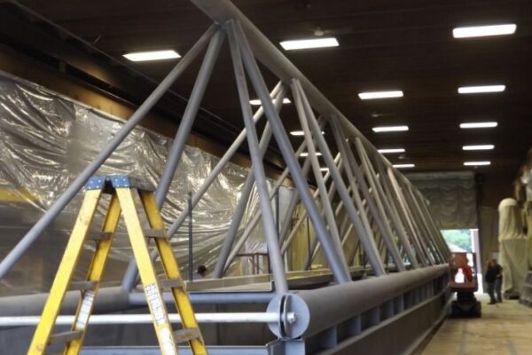 mid-city-steel-mid-city-steel-milwaukee-intermodal-station-special-project-gallery-10 A steel truss bridge structure indoors, with a yellow ladder in front and plastic sheets covering the walls.