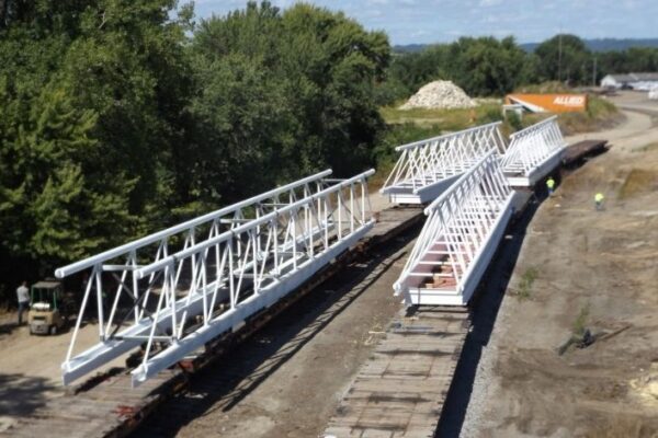 mid-city-steel-mid-city-steel-milwaukee-intermodal-station-special-project-gallery-3 Two large white steel bridge sections rest on flatcars beside a dirt road, surrounded by trees and construction materials.