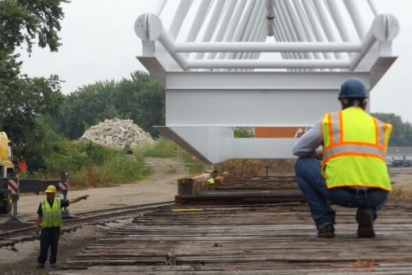 mid-city-steel-mid-city-steel-milwaukee-intermodal-station-special-project-gallery-8 Two workers guide a large metal structure being placed over railroad tracks; one crouches, one directs.