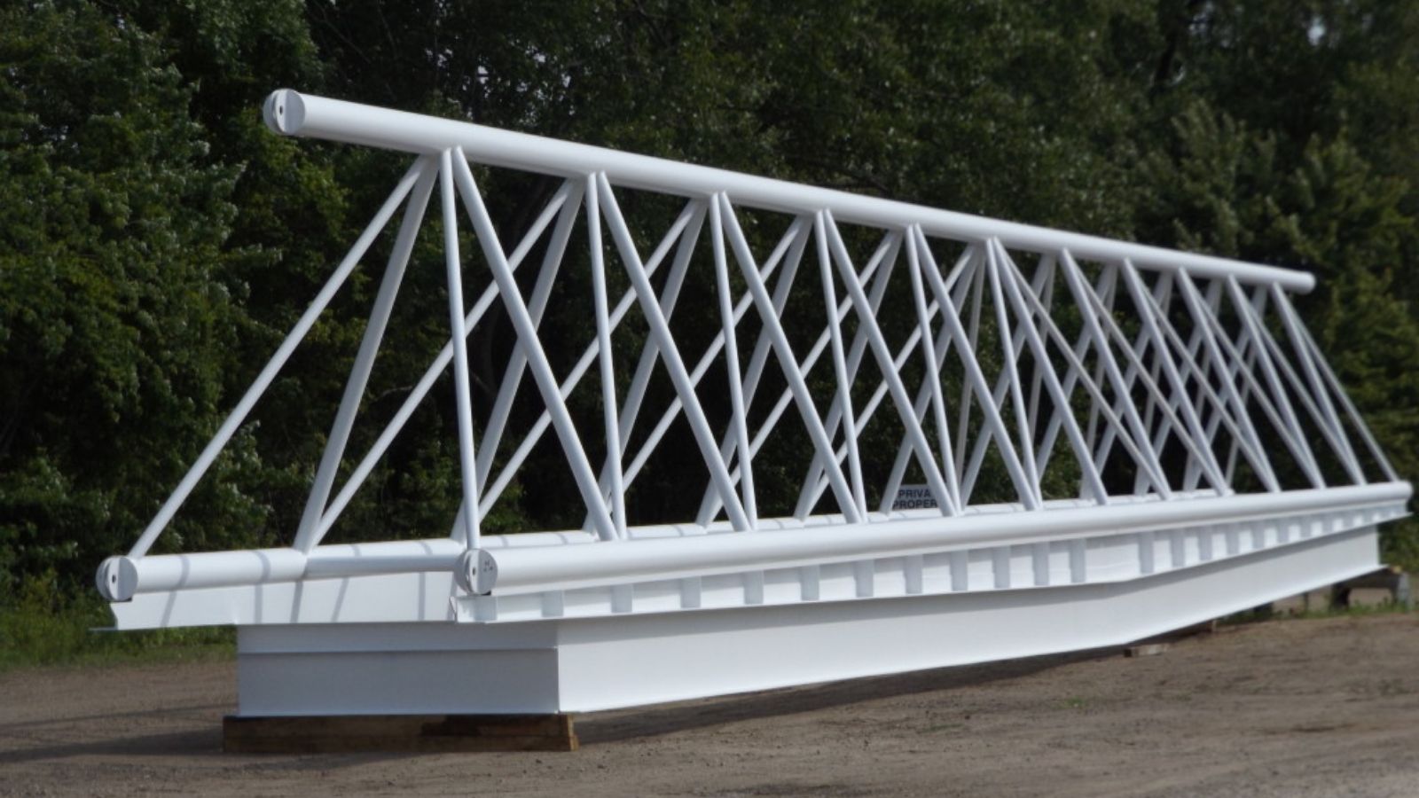 A large, white metal truss bridge section is resting on the ground, surrounded by green trees and grass.