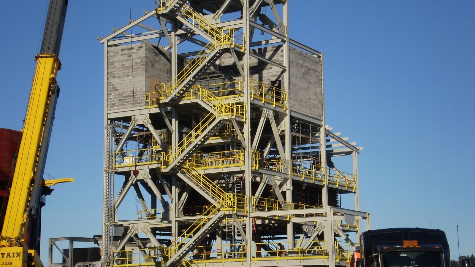 Steel industrial structure with yellow staircases and platforms against a clear blue sky, with vehicles nearby.
