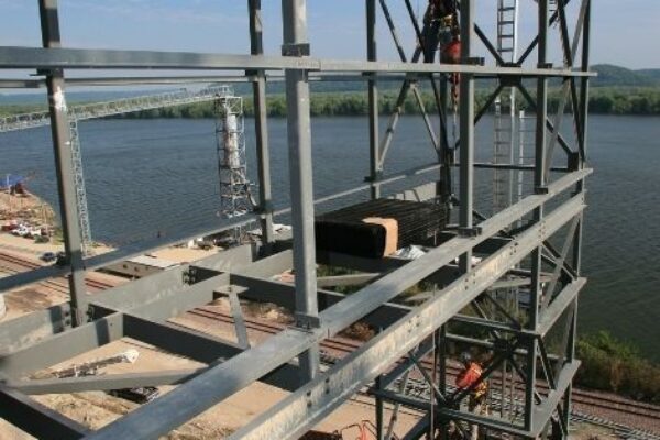 mid-city-steel-pattison-frac-sand-plant-industrial-project-gallery-7 A worker stands on a steel framework by a river, with construction equipment below and blue sky above.