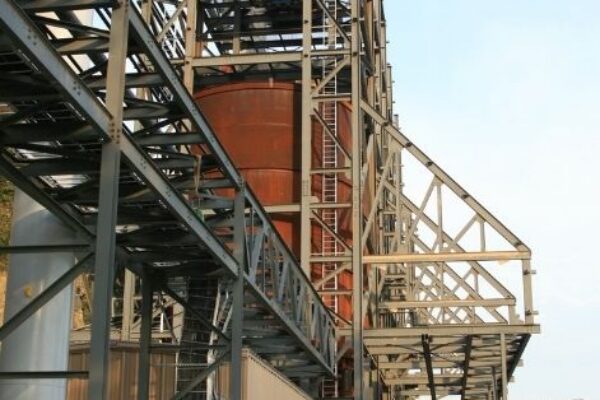 mid-city-steel-pattison-frac-sand-plant-industrial-project-gallery-9 Steel framework of an industrial building under construction, with two people walking nearby on a dirt path.