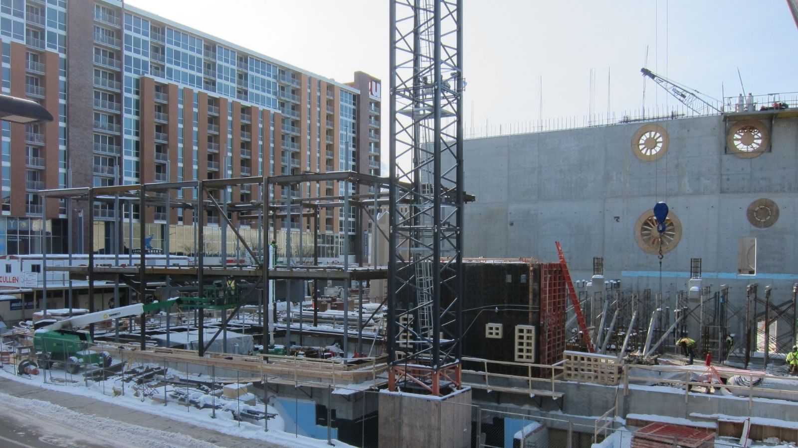 Construction site with steel framework, concrete walls, cranes, and workers; apartment building in the background.