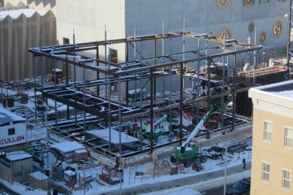 Steel beams form the skeleton of a building under construction on a snowy city lot, with workers and equipment onsite.