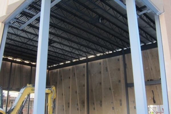 Large open warehouse under construction with exposed steel beams and a yellow excavator inside.