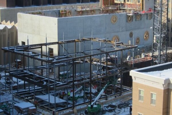 Steel framework and construction equipment at a snowy building site next to partially completed concrete and finished buildings.