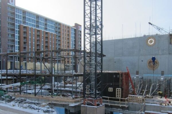 Snowy construction site with steel framework, crane, and tall apartment building in the background.