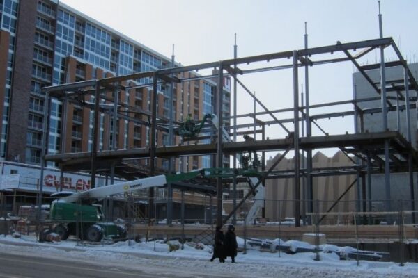 Steel framework of a building under construction, with cranes and workers, on a snowy urban street.