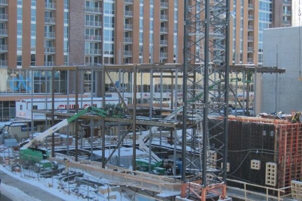 Construction site with steel framework, cranes, and workers; snow on the ground and tall buildings in the background.