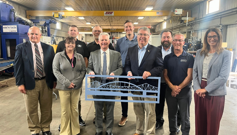 Senator Brad Pfaff, Wisconsin Economic Development Corporation and La Crosse Area Chamber of Commerce representatives, and local business leaders gather inside Mid-City Steel for a roundtable discussion, with a metal bridge cutout displayed in front.